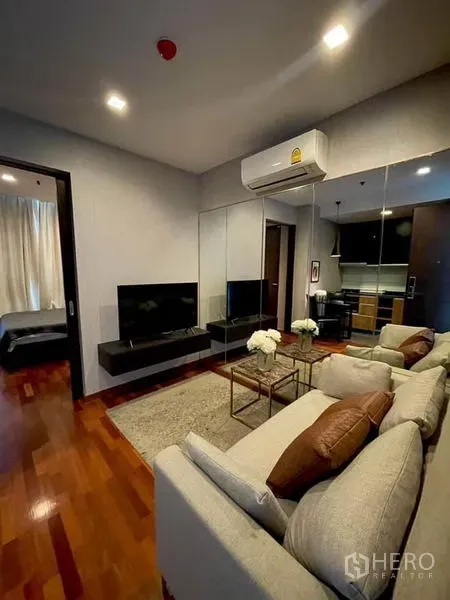 Condominium for rent or sale in Ratchathewi, Bangkok - Modern living room with gray sofa, wall-mounted TV, mirrored wall, and warm wood floors beside the open kitchen.