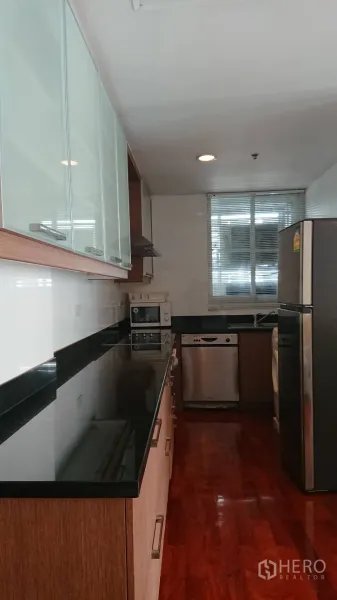 Condominium for rent in Pathum Wan, Bangkok - Galley kitchen with black granite countertops, glass-front cabinets, stainless refrigerator, dishwasher and microwave by a window.