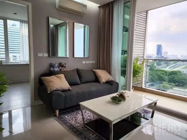 Condominium for rent in Huai Khwang, Bangkok - Daylit lounge with gray sofa beside full-height windows and city outlook.