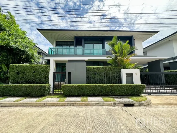 บ้าน ให้เช่าหรือขายในBang Kapi, Bangkok - หน้าบ้านเดี่ยวสองชั้นสมัยใหม่ ระเบียงกระจก พุ่มไม้ตัดแต่ง และรั้วหน้าบ้านท่ามกลางท้องฟ้าโปร่ง.