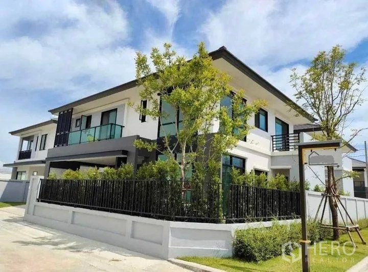 Detached House for rent in Bang Bo, Samut Prakan - Modern two-storey detached house on a corner plot with black fence and lush greenery under a blue sky.
