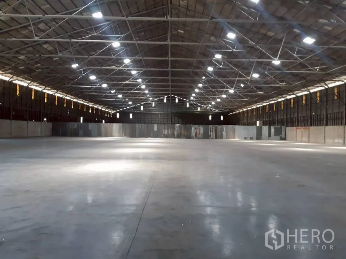Warehouse for rent in Pak Kret, Nonthaburi - Symmetrical view down a vast warehouse lit by ceiling fixtures across a smooth concrete floor.