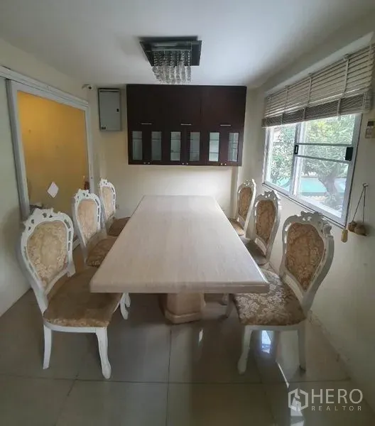 Townhouse for rent in Phaya Thai, Bangkok - Dining room with a long table and ornate chairs, wall cabinets, window blinds and a modern light fixture.