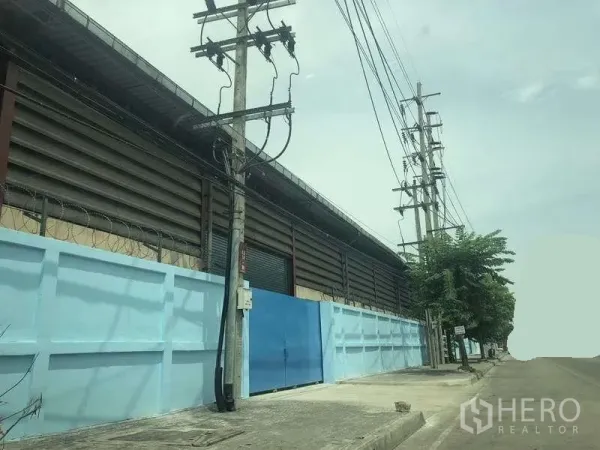 Warehouse for rent in Phra Pradaeng, Samut Prakan - Street view of a secure warehouse exterior with blue perimeter wall, metal gate and power lines along a wide road.