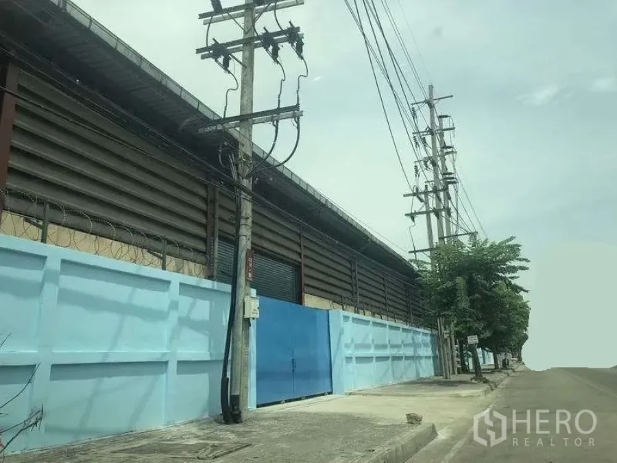 Warehouse for rent in Phra Pradaeng, Samut Prakan - Street view of a secure warehouse exterior with blue perimeter wall, metal gate and power lines along a wide road.