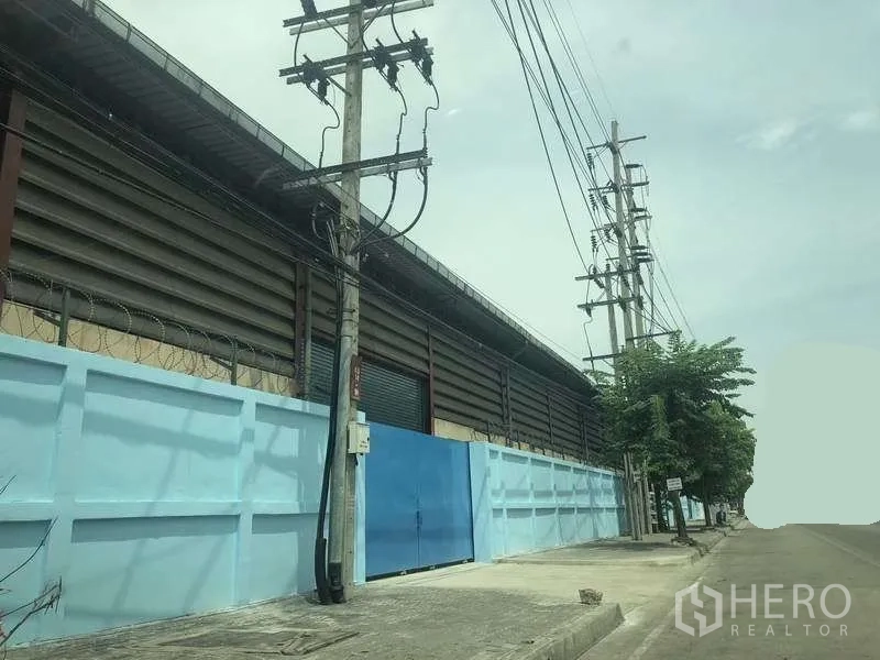 Warehouse for rent in Phra Pradaeng, Samut Prakan - Street view of a secure warehouse exterior with blue perimeter wall, metal gate and power lines along a wide road.