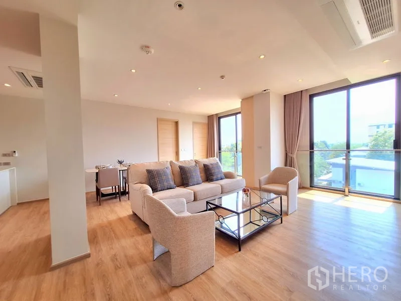 อพาร์ทเมนท์ ให้เช่าในBang Na, Bangkok - ห้องนั่งเล่นแบบโอเพ่นที่สว่าง มีโซฟาชุด พื้นไม้ และหน้าต่างสูงจรดเพดานเชื่อมระเบียง
