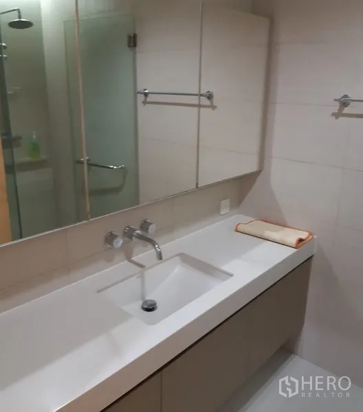 Condominium for rent in Khlong Toei, Bangkok - Contemporary bathroom with long vanity, under-mount sink, mirrored cabinets and a glass-enclosed shower.