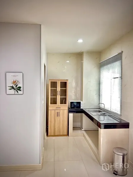 Townhouse for rent in Bang Kapi, Bangkok - Compact light-toned kitchen with window blinds, stainless sink, microwave and wood pantry cabinet.