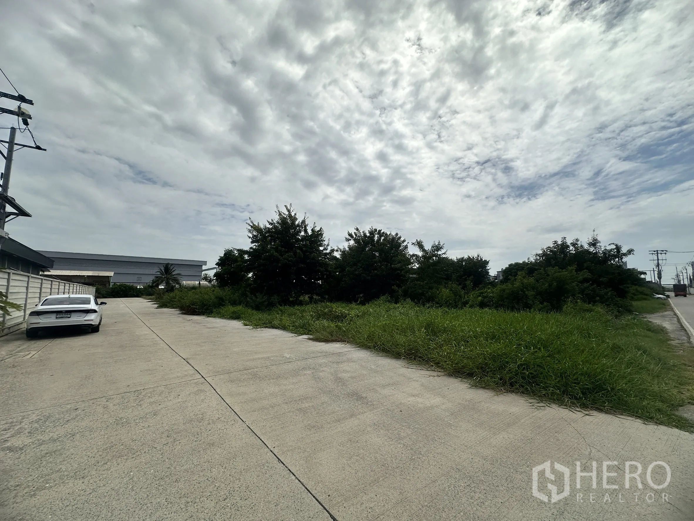 Land for sale in Sai Noi, Nonthaburi - Concrete access road beside a boundary wall and greenery, with nearby warehouse buildings.