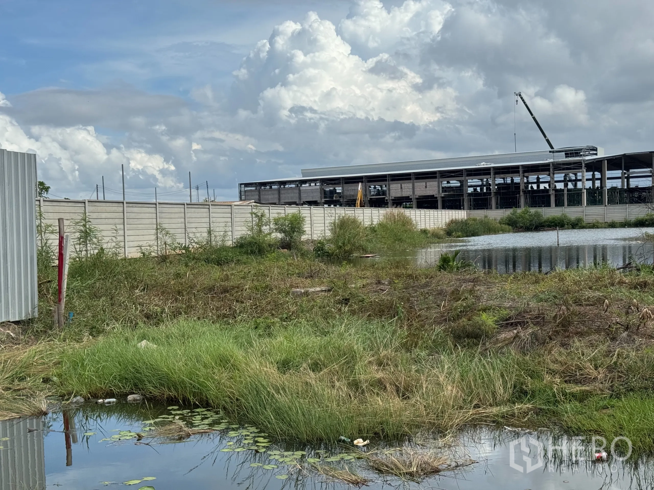 Land for sale in Samut Prakan - Vacant land with grass and a drainage pond beside a boundary wall near warehouses in Phra Samut Chedi.