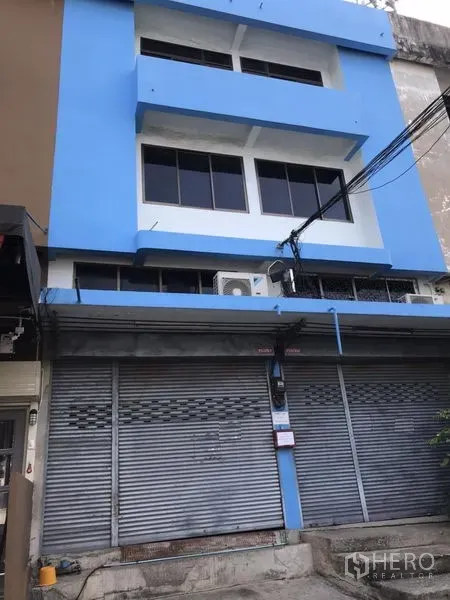 店屋 出售在Suan Luang, Bangkok - 三层蓝色商铺外立面，一层金属卷闸门关闭，楼上大窗与室外空调机组，位于城市小巷。