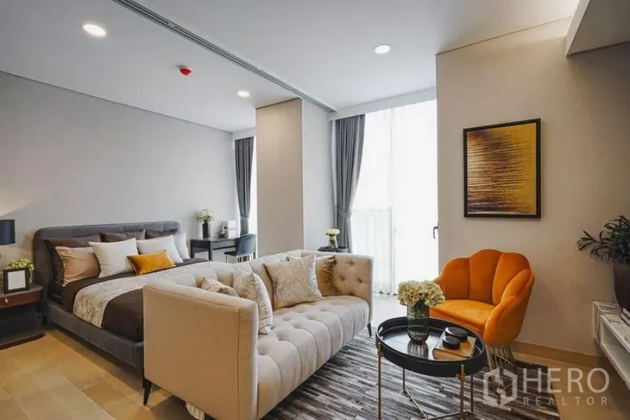คอนโด ขายในKhlong Toei, Bangkok - ห้องนอนเชื่อมต่อห้องนั่งเล่น มีหน้าต่างเต็มบาน โซฟาสีเบจ และเก้าอี้สีส้มเด่น