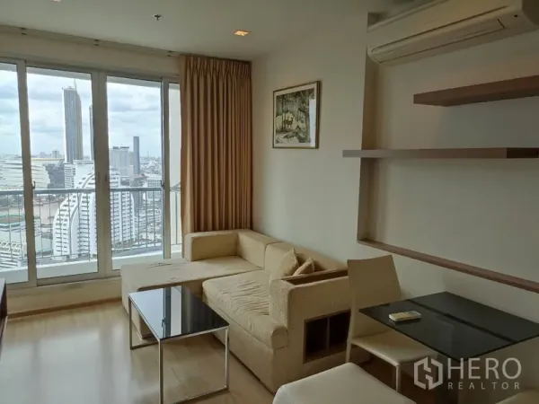Condominium for rent or sale in Sathon, Bangkok - Living area with corner sofa, two-seat dining table and floor-to-ceiling windows with city views.