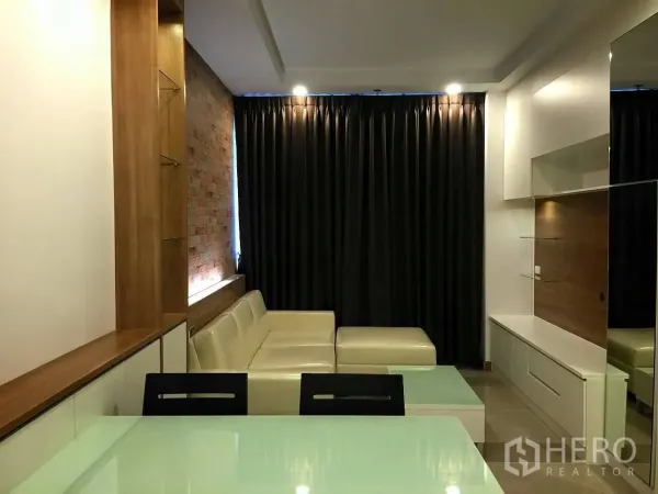 Condominium for sale in Huai Khwang, Bangkok - Modern living room with an L-shaped cream sofa, brick feature wall, built-in TV cabinet and blackout curtains in a Huai Khwang condo.