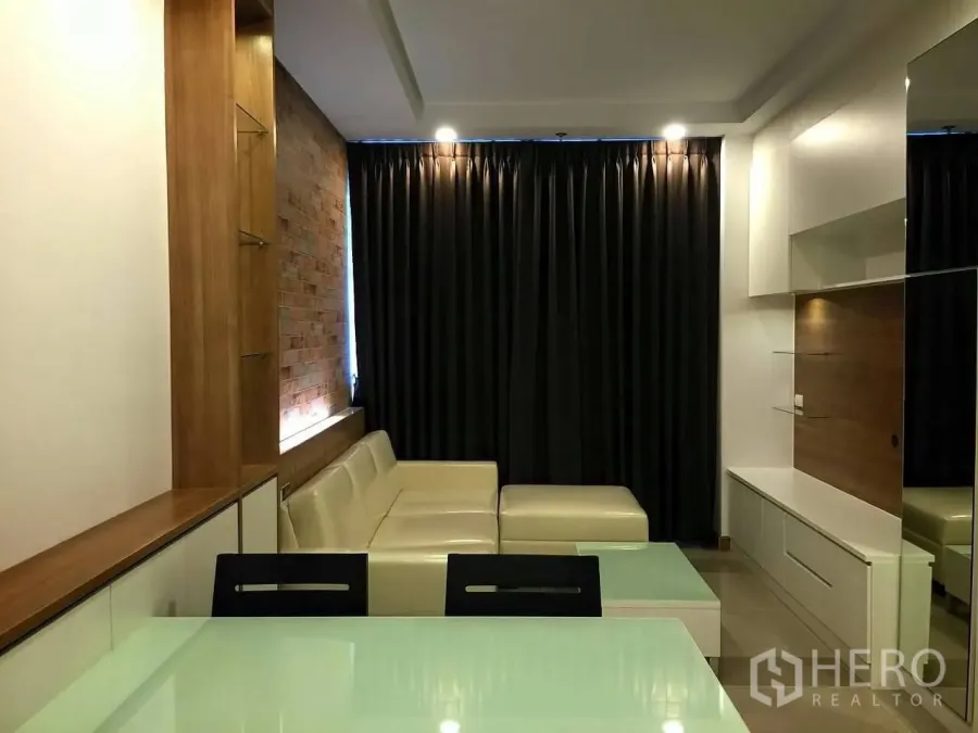 Condominium for sale in Huai Khwang, Bangkok - Modern living room with an L-shaped cream sofa, brick feature wall, built-in TV cabinet and blackout curtains in a Huai Khwang condo.