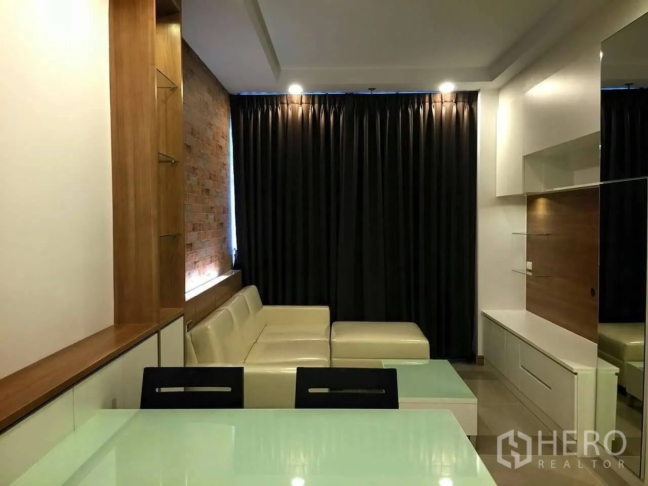 Condominium for sale in Huai Khwang, Bangkok - Modern living room with an L-shaped cream sofa, brick feature wall, built-in TV cabinet and blackout curtains in a Huai Khwang condo.