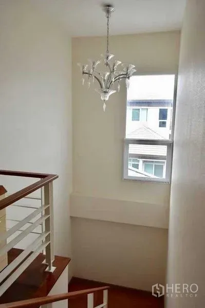 Detached House for sale in Bueng Kum, Bangkok - Stairwell with window and contemporary chandelier above a wooden handrail and white walls.