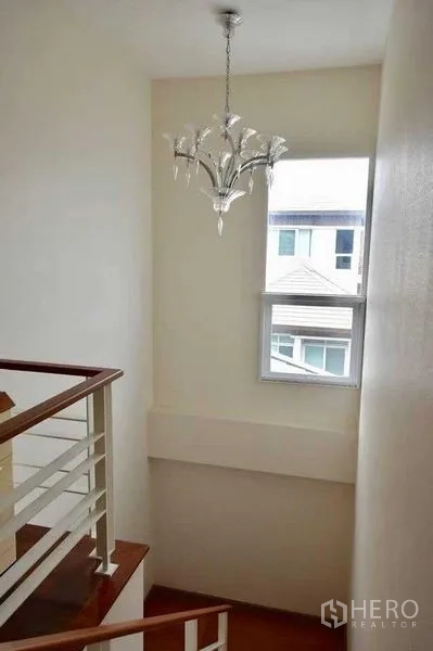 Detached House for sale in Bueng Kum, Bangkok - Stairwell with window and contemporary chandelier above a wooden handrail and white walls.