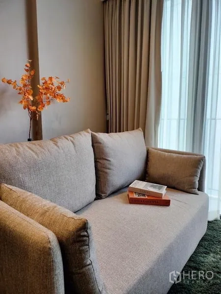 อพาร์ทเมนท์ ให้เช่าหรือขาย - ห้องนั่งเล่นโทนอุ่นพร้อมโซฟาสีเทา หนังสือ แจกันใบไม้สีส้ม และผ้าม่านสูงจรดเพดาน