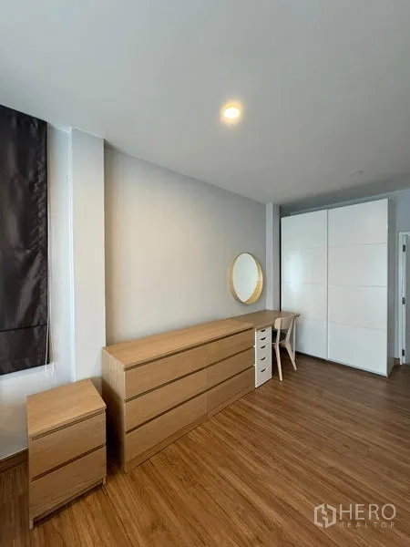 Detached House for rent in Don Mueang, Bangkok - Bedroom storage area with long wooden dresser, round mirror and white wardrobe on wood floors.