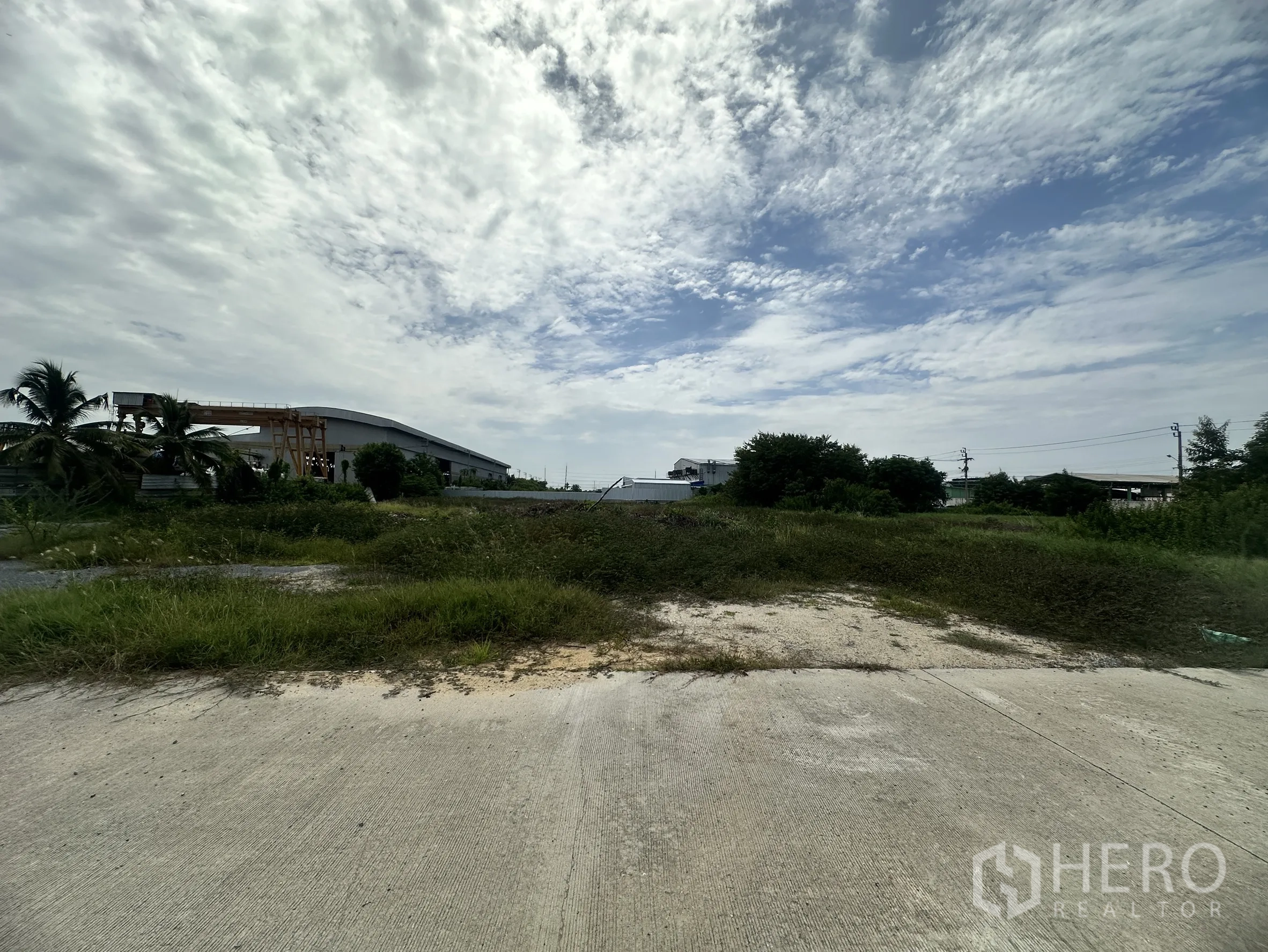 Land for sale in Sai Noi, Nonthaburi - Vacant plot with grass and shrubs viewed from a concrete road, warehouses in the distance and utility poles.