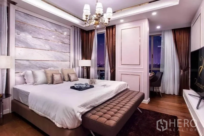 Condominium for rent in Huai Khwang, Bangkok - Master bedroom with chandelier, corner windows, TV and upholstered bench.
