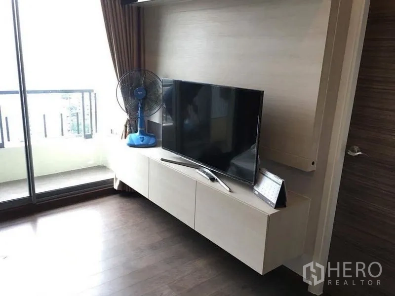 Condominium for rent in Ratchathewi, Bangkok - Bright living area with wall-mounted TV unit and sliding glass door to a balcony.