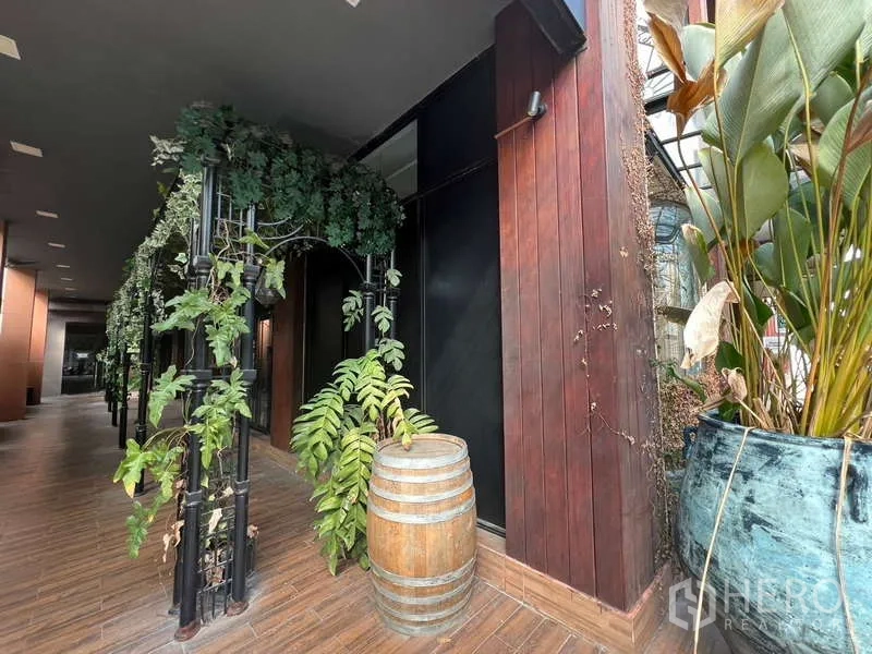 零售空间 出租在Watthana, Bangkok - 建筑侧立面为木饰面与缠藤铁艺拱架，旁置装饰酒桶与花盆。