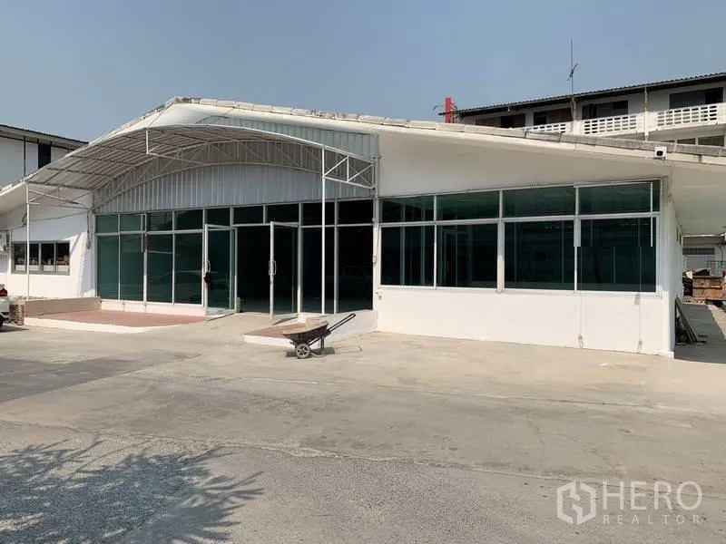 货栈 出租在Mueang Pathum Thani, Pathum Thani - 仓库外立面为白色墙体与整面玻璃，钢制雨棚，前方宽阔混凝土场地可停车。