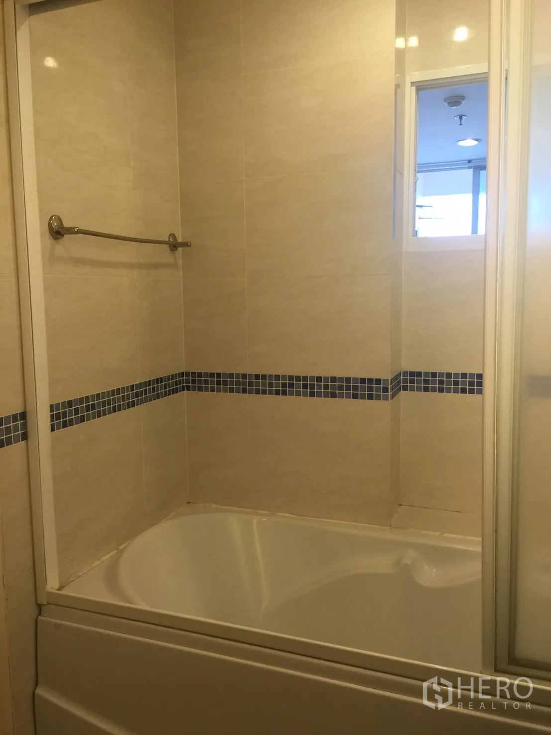 Condominium for rent in Watthana, Bangkok - Bathroom with built-in bathtub, glass partition, and beige tiles accented by a blue mosaic strip.
