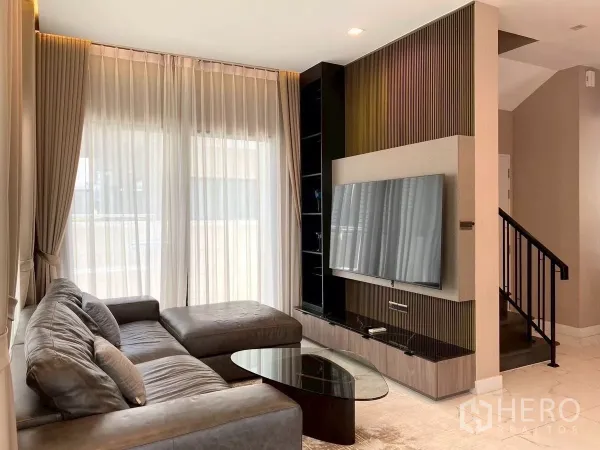 Detached House for rent in Bang Phli, Samut Prakan - Cozy living room with L-shaped leather sofa, wall-mounted TV unit and staircase.
