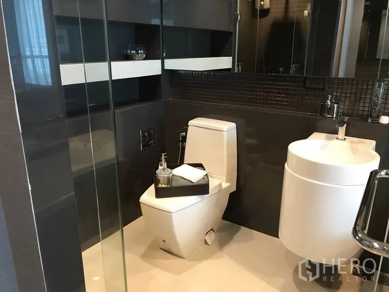 公寓 出租在Sathon, Bangkok - 现代卫浴，设玻璃淋浴间、方形坐便和圆形面盆，深色墙砖点缀。