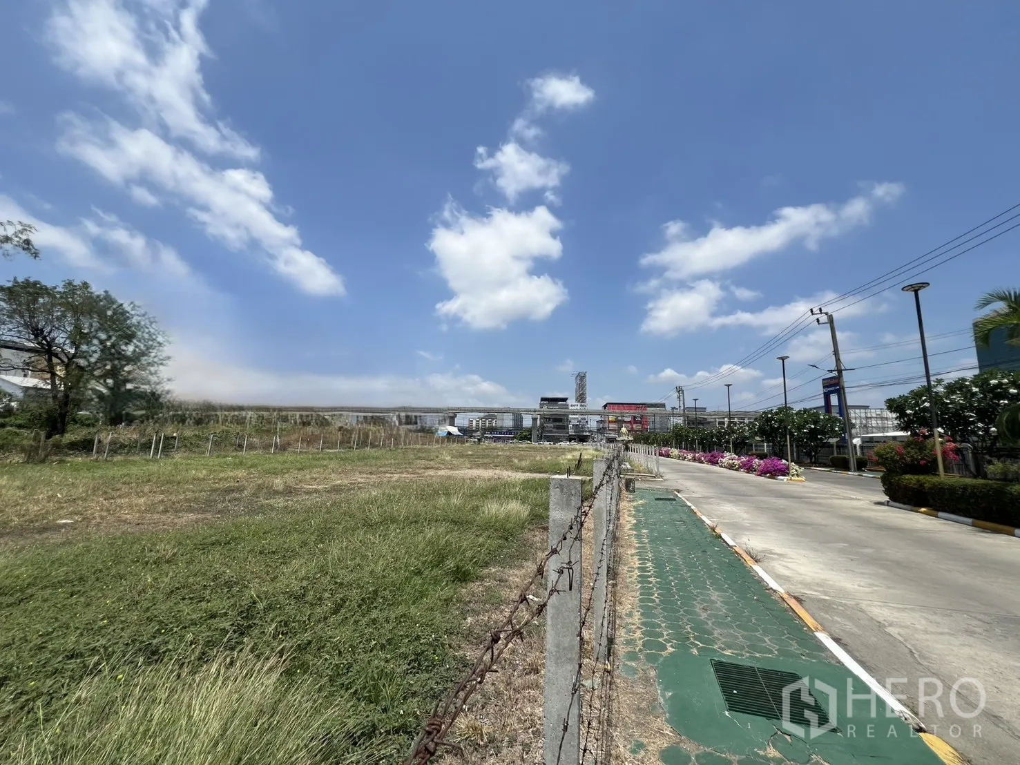 Land for rent in Mueang Samut Prakan, Samut Prakan - Vacant grassy land plot bordered by posts and barbed wire beside a paved road with streetlights and landscaped verge.