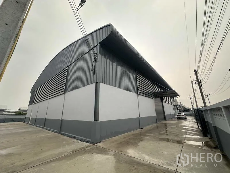 Factory for rent in Pathum Thani - Side view of metal-clad warehouse walls with service lane, perimeter fence and power lines.