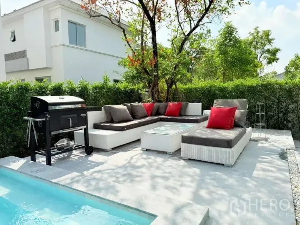 Detached House for rent or sale in Bang Phli, Samut Prakan - Poolside patio featuring an outdoor sofa set with red cushions and a barbecue grill shaded by trees.