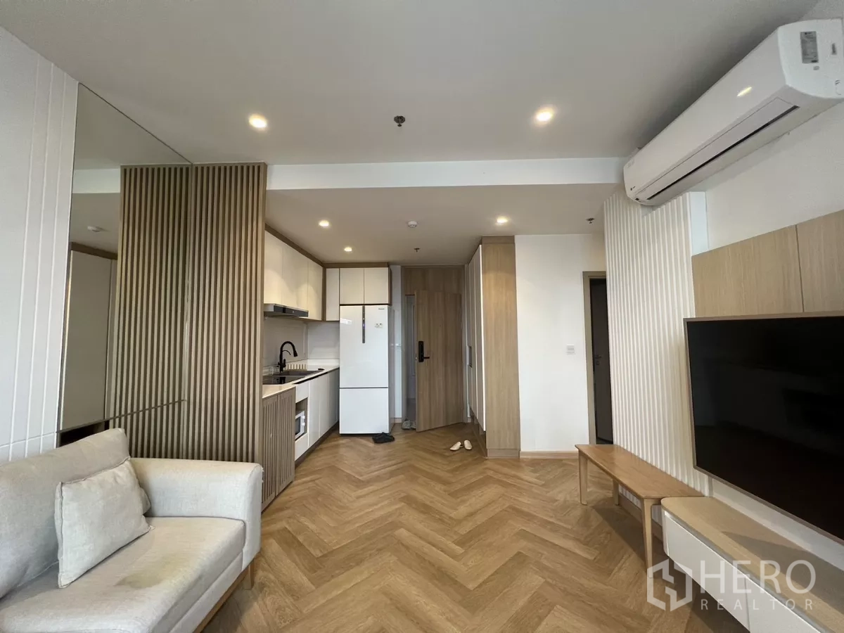 Condominium for rent or sale in Chatuchak, Bangkok - Open‑plan view toward entry and kitchenette with air‑conditioner, tall fridge and light wood accents.