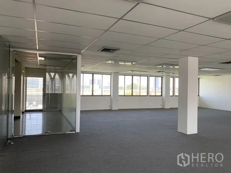 Office Space for rent in Bang Kapi, Bangkok - Expansive office space with support columns, suspended ceiling lighting, and an adjacent glass-walled room beside wide windows.