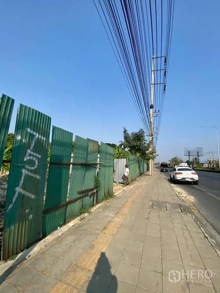 Land for sale in Bang Khae, Bangkok - Main road frontage with sidewalk, green site fence and overhead power lines beside the land.