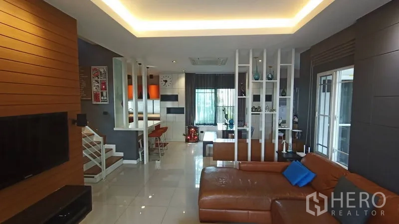 独立式住宅 出售在Prawet, Bangkok - 宽敞客厅，L形皮沙发，通透隔断连接餐区与楼梯。