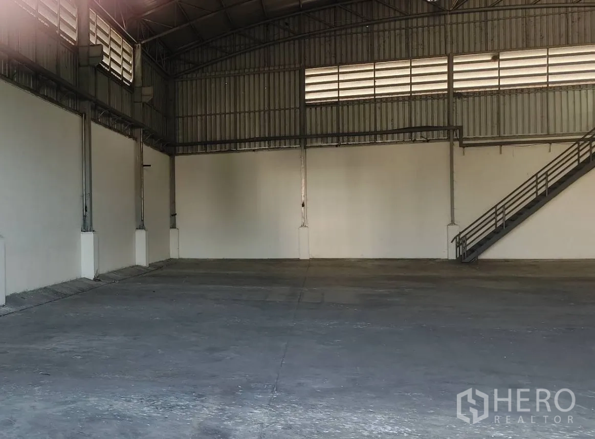工厂 出租在Lat Lum Kaeo, Pathum Thani - 开阔仓储地面，高钢结构屋顶与白色墙面，侧边楼梯通往办公室。