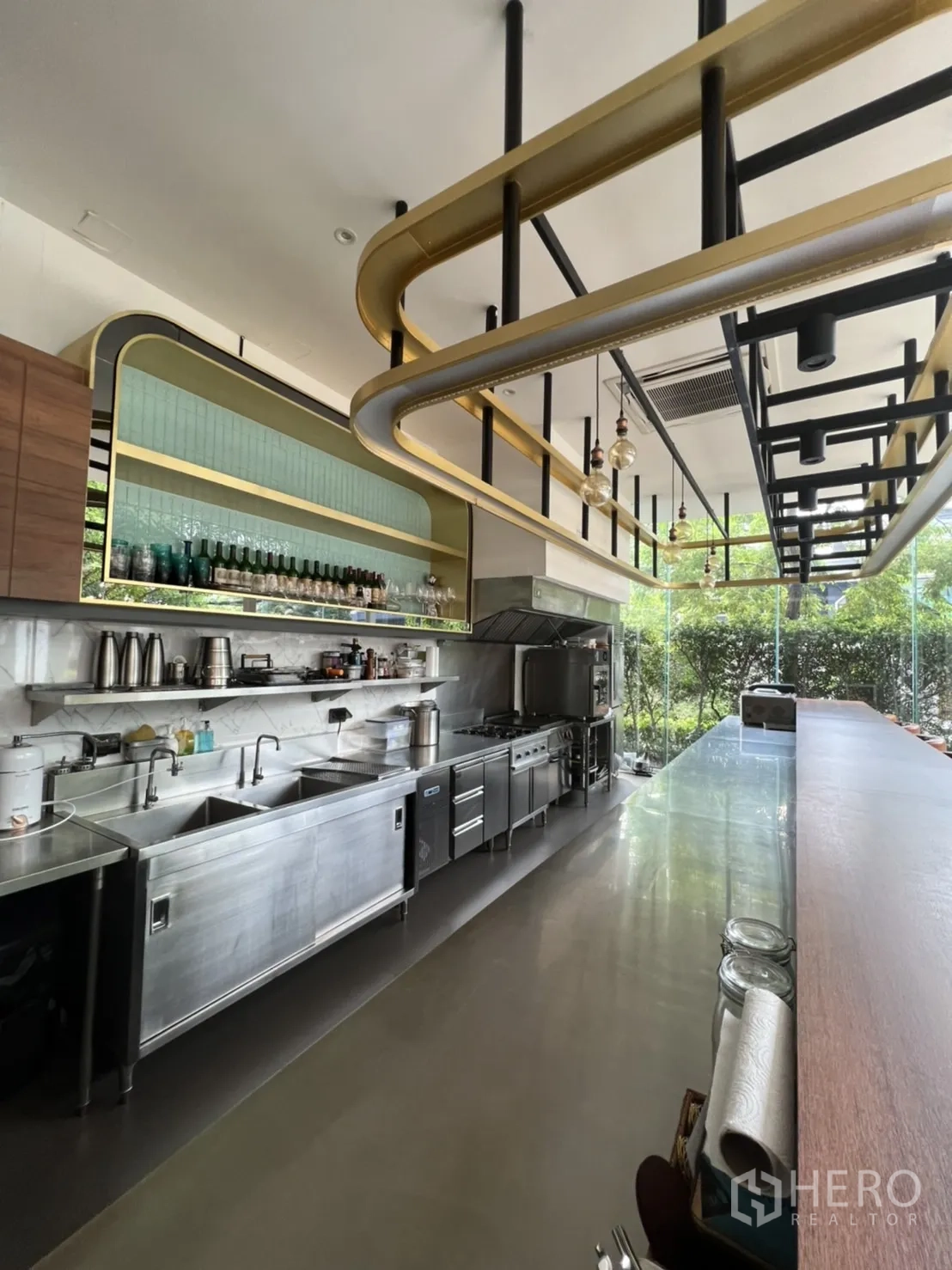 พื้นที่ขายปลีก ให้เช่าในWatthana, Bangkok - ครัวเชิงพาณิชย์สแตนเลสครบชุด พร้อมอ่างล้างมือ เตาอบ เตาแก๊ส และชั้นวาง ใต้โครงไฟสีทอง เห็นสวนด้านนอก