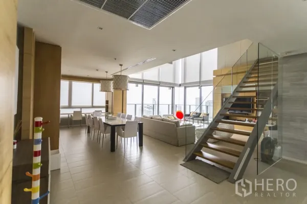 Condominium for rent in Bang Lamung, Chon Buri - Spacious duplex living and dining room with glass staircase and city-sea panorama.