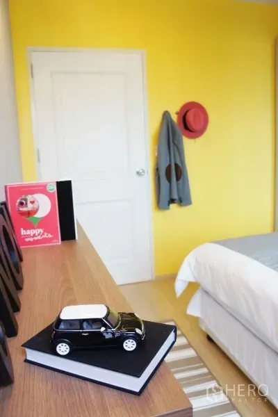 คอนโด ให้เช่าในPhaya Thai, Bangkok - มุมห้องนอนผนังสีเหลือง ตู้ลิ้นชัก ของตกแต่ง และขอบเตียงข้างประตูสีขาว