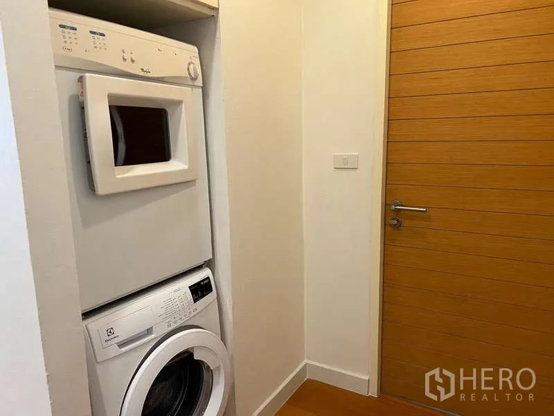 Condominium for rent in Bang Kho Laem, Bangkok - Laundry nook with stacked washer and dryer next to wooden door.