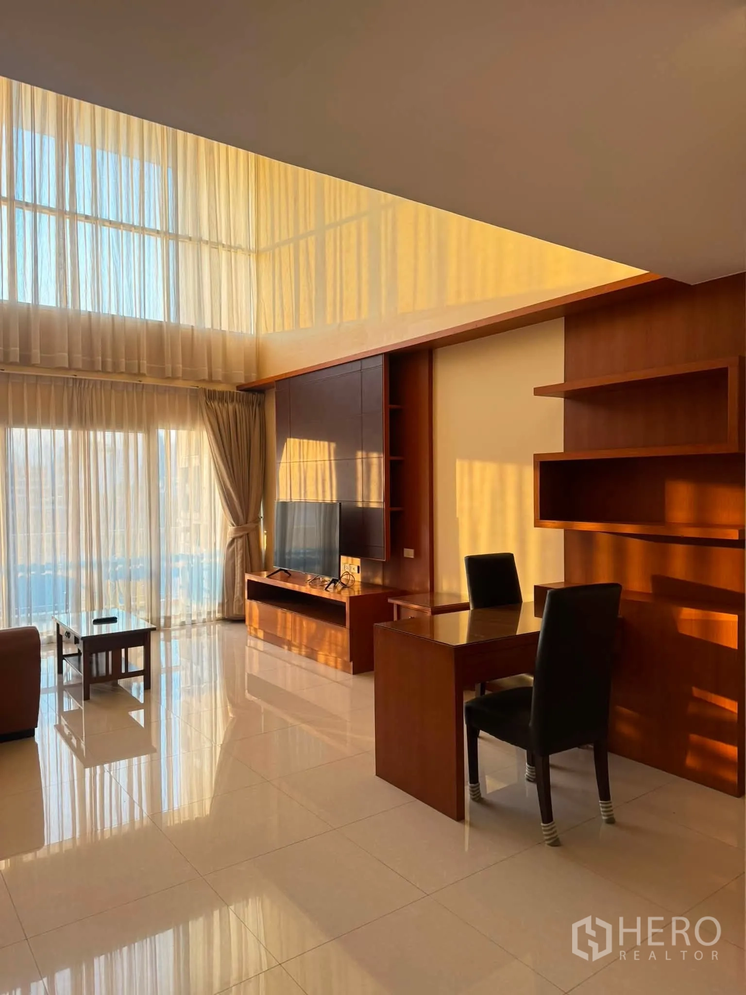 Condominium for rent in Khlong Toei, Bangkok - Bright living room with floor-to-ceiling curtains, work desk, wall-mounted TV, and wooden shelving.