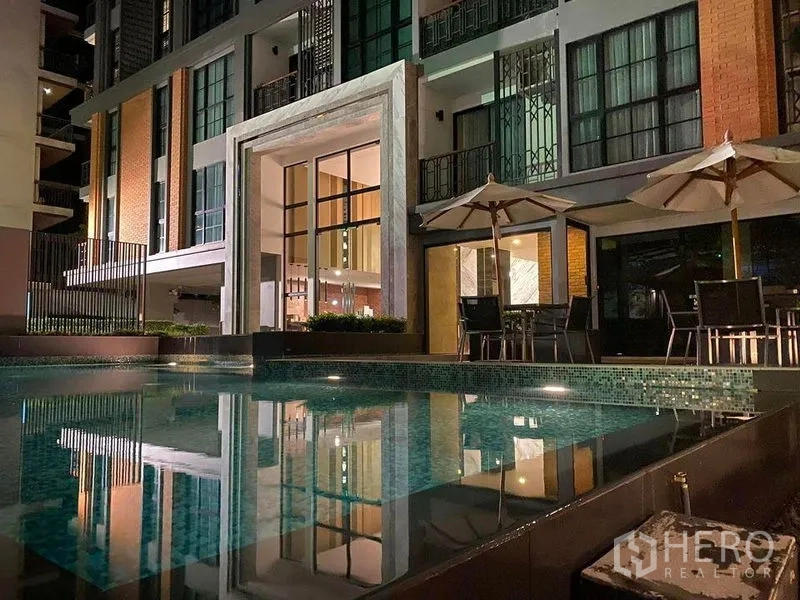 公寓 出租在Pathum Wan, Bangkok - 夜色下的公寓外立面与点亮的泳池露台和遮阳伞。