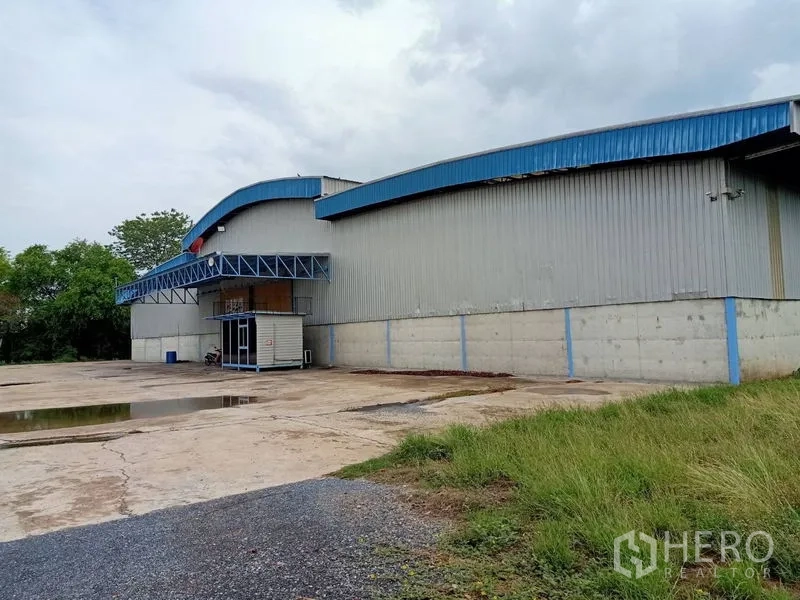 Factory for rent in Chaloem Phra Kiat, Saraburi - Metal-clad factory exterior with blue roofline, loading canopy and wide concrete apron beside greenery.