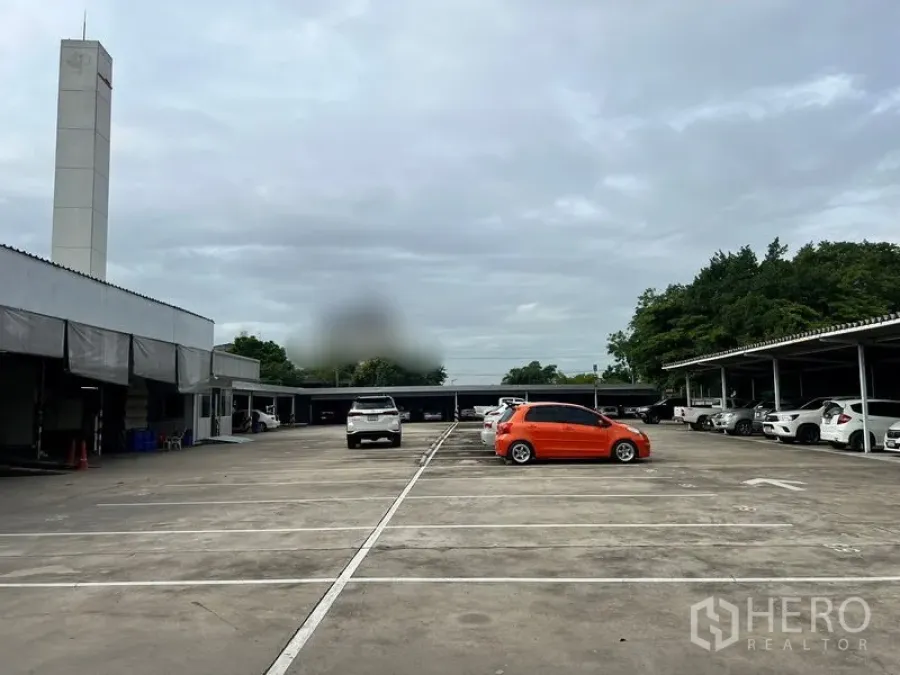 ที่ดิน ให้เช่าในBang Na, Bangkok - มุมมองกึ่งกลางลานปูนขนาดใหญ่ มีโรงจอดรถสองฝั่งและแนวต้นไม้ด้านหลัง