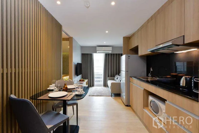 Apartment for rent in Khlong Toei, Bangkok - Long galley kitchen with built-in appliances opposite a dining table, opening to lounge and balcony.